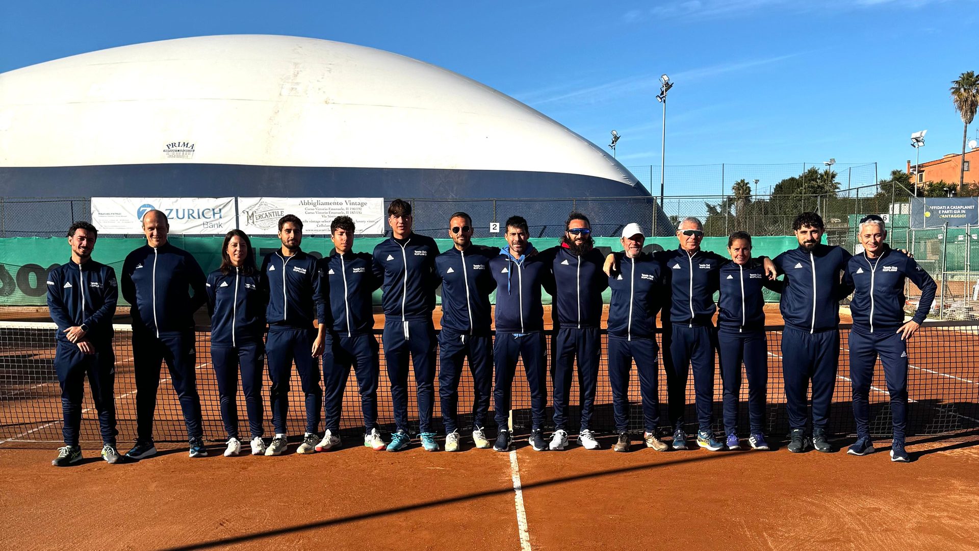 Lo staff della scuola tennis schierato sulla terra battuta, davanti al pallone del campo coperto